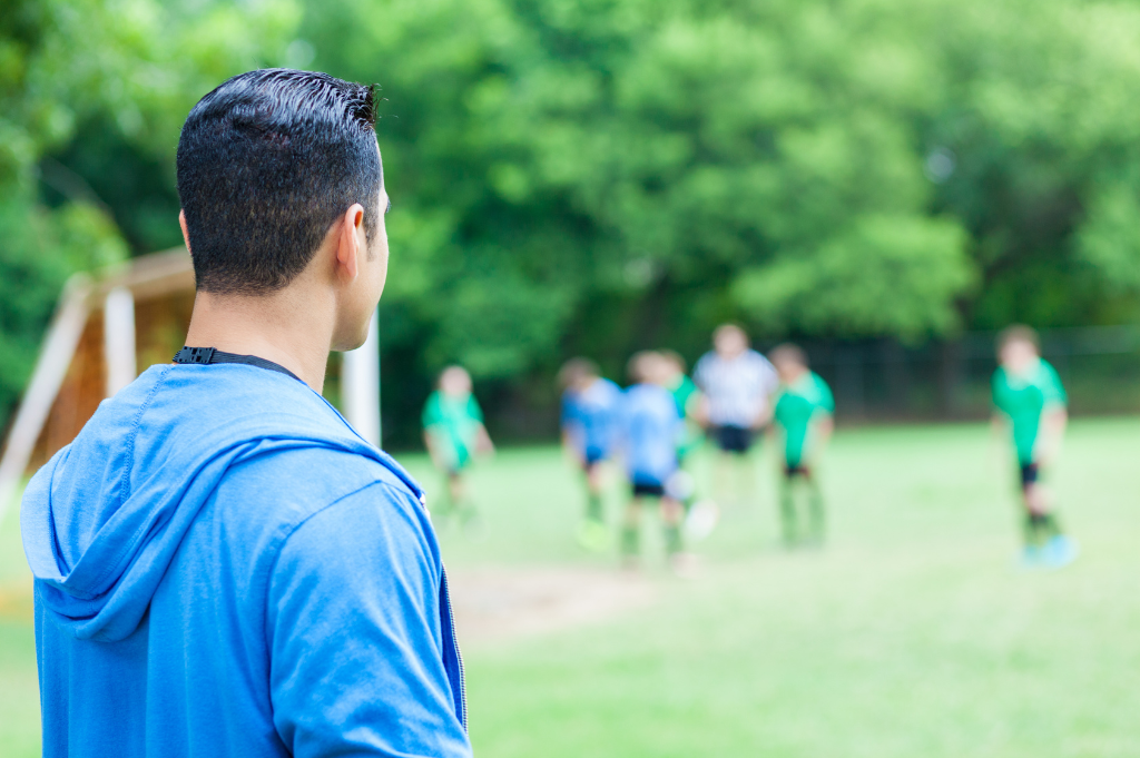 A football scout is watching grassroots players from the sidelines. He is paying attention to what football scouts look for at the grassroots level.
